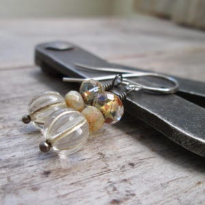 May include: Close-up of handmade earrings featuring clear glass beads with silver stripes and faceted, iridescent beads. The earrings are on a silver hook and are set against a blurred background of a metal tool and a wooden surface.
