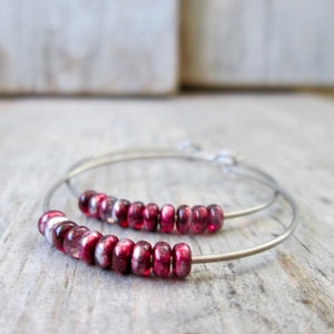 May include: A pair of silver hoop earrings with a row of deep red beads. The beads are evenly spaced along the bottom half of the hoops. The earrings are displayed on a wooden surface.