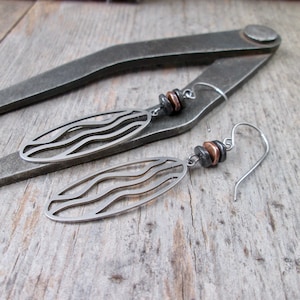 May include: A pair of silver earrings with a wavy design and copper beads. The earrings are hanging from a metal tool.