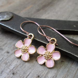 May include: A pair of flower earrings. Each earring features a pink enamel flower with a gold-colored center and a gold-colored border. The earrings have copper-colored ear wires and are displayed on a wooden surface.