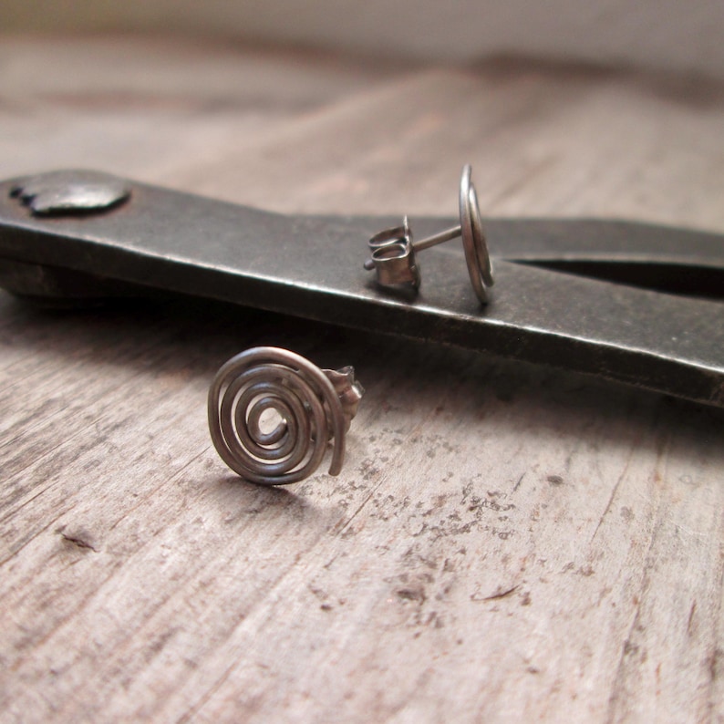 May include: A pair of silver-toned spiral stud earrings. One earring is in focus, displaying a coiled design. The other earring is slightly out of focus, showing the post and backing. The earrings are on a wooden surface.