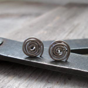 May include: A pair of silver-toned spiral stud earrings. Each earring features a coiled design, creating a circular shape. The earrings are displayed on a dark gray surface, with a wooden background.