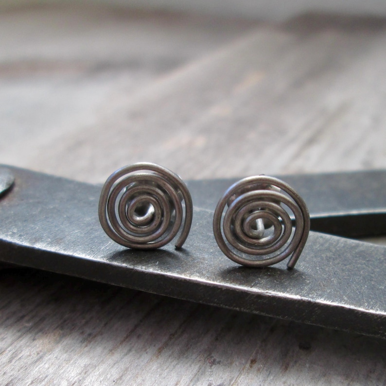 May include: A pair of silver-toned spiral stud earrings. Each earring features a coiled design, creating a circular shape. The earrings are displayed on a dark gray surface, with a wooden background.