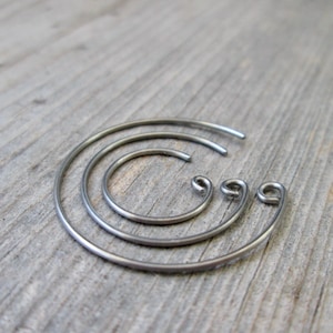 May include: Three silver-colored, spiral-shaped earrings of varying sizes are arranged on a wooden surface. The earrings have a simple, minimalist design, with a smooth, metallic finish. The largest earring is a complete spiral, while the others are partial.