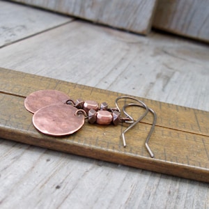 May include: A pair of copper earrings with hammered round discs and copper beads. The earrings are hanging from a wooden ruler.