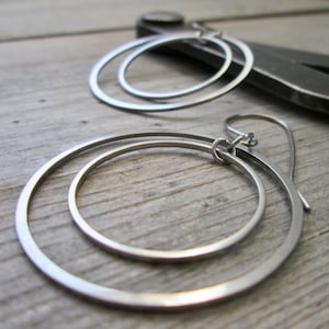 May include: Silver hoop earrings with a double-circle design. The earrings are made of a shiny, metallic material and have a simple, modern aesthetic. The earrings are shown on a wooden surface.
