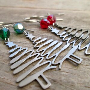 May include: Silver-toned Christmas tree earrings with dangling red and green faceted beads. The earrings are made of a cut-out design in the shape of a Christmas tree with a star at the top. The earrings are displayed on a wooden surface.