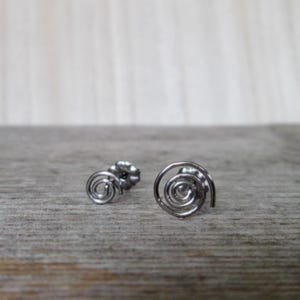 May include: A pair of silver-colored spiral stud earrings. The earrings are made of thin, coiled metal, with a post and backing visible on one earring. The earrings are displayed on a light brown wooden surface.