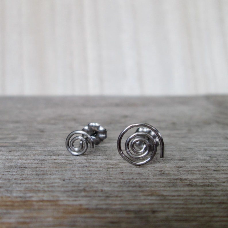 May include: A pair of silver-colored spiral stud earrings. The earrings are made of thin, coiled metal, with a post and backing visible on one earring. The earrings are displayed on a light brown wooden surface.