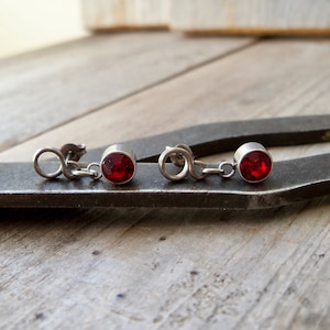 May include: A pair of silver earrings with red gemstone accents. The earrings feature a small chain connecting the gemstone to the earring post.