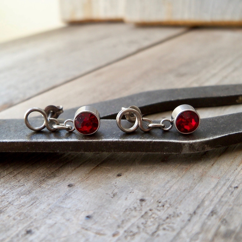 May include: A pair of silver earrings with red gemstone accents. The earrings feature a small chain connecting the gemstone to the earring post.