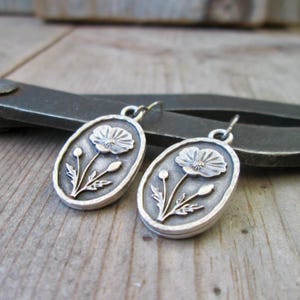 May include: A pair of silver-toned oval earrings. Each earring features a detailed poppy flower design with a stem, leaves, and buds. The earrings are set against a wooden surface with a metal tool in the background.
