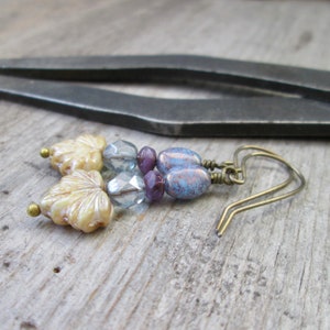 May include: A pair of earrings with a blue and purple color scheme. The earrings feature a leaf-shaped charm, blue and purple glass beads, and a blue and white speckled oval bead.