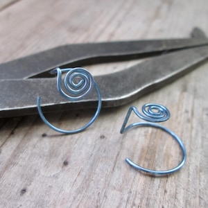 May include: Two blue spiral earrings. Each earring features a coiled spiral design at the top, with a curved, open bottom. The earrings are made of a thin, metallic material. The background includes a wooden surface and a metal tool.