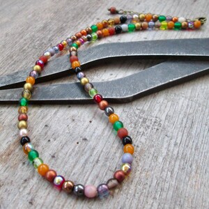 May include: A colorful beaded necklace with a variety of small, round beads in shades of red, orange, yellow, green, and purple. The necklace is displayed on a wooden surface with a metal tool in the background.