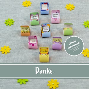 Puede incluir: Nueve etiquetas de regalo de papel de colores con diferentes mensajes de agradecimiento en alemán. Las etiquetas están decoradas con flores y están dispuestas sobre un fondo de tela gris claro. El texto en las etiquetas dice: "Danke schon", "Kleiner Dank", "Süßer Dank", "Danke" y "Direkt Download".