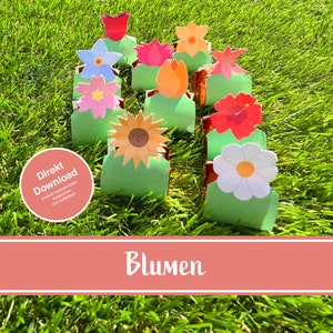 Pode incluir: Um conjunto de caixas imprimíveis em forma de flor, com uma variedade de cores e designs, incluindo um girassol, uma margarida, um hibisco e uma tulipa. As caixas estão dispostas em uma fila sobre uma superfície verde coberta de grama. O texto "Direkt Download" é visível em um círculo no lado esquerdo da imagem. O texto "Blumen" é visível em uma faixa na parte inferior da imagem.