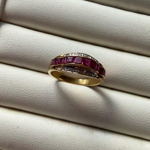 May include: A gold ring featuring a central row of five square-cut deep red rubies flanked by two rows of small, clear round-cut diamonds. The band is polished yellow gold, showcasing a multi-stone channel setting that creates a layered appearance.