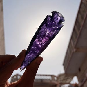 Può includere: Una mano tiene in mano un oggetto traslucido a forma di punta di freccia viola contro un cielo luminoso. L'oggetto ha una superficie strutturata e un bordo scuro. La luce brilla attraverso l'oggetto, evidenziandone il colore e la forma.