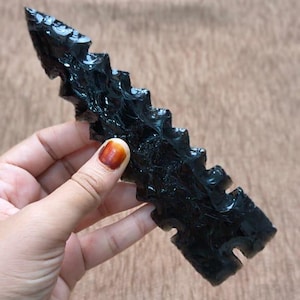 Puede incluir: Una punta de flecha de obsidiana negra con borde dentado. La punta de flecha se sostiene en una mano, mostrando su superficie oscura y brillante. El fondo es una tela texturizada de tono neutro.