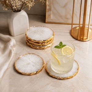 Peut inclure: Un ensemble de dessous de verre ronds en résine blanche avec des bords irréguliers peints en doré. Un dessous de verre soutient un verre cannelé transparent contenant une boisson au citron, tandis qu'une pile de trois autres repose à côté.