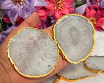 May include: White and gray agate coasters with gold-painted edges. The coasters have a natural, organic shape and are displayed against a backdrop of colorful flowers. These decorative coasters are ideal for protecting surfaces.