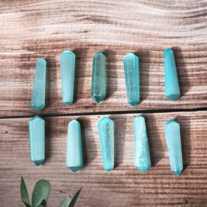 May include: Ten light blue crystal wands are arranged on a wooden surface. The wands have a hexagonal shape and pointed tips. The crystals vary slightly in color and clarity, with some appearing more translucent than others. The background is a warm-toned wood.