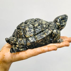 May include: A decorative turtle figurine with a mosaic-like shell pattern in shades of gray, black, and gold. The turtle is resting on a hand, showcasing its detailed design and craftsmanship. A unique home decor item.