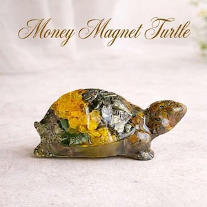 May include: A decorative turtle figurine with a shell filled with yellow and green elements. The turtle is brown and green with the text "Money Magnet Turtle" in gold script above it. The figurine is on a light-colored surface.
