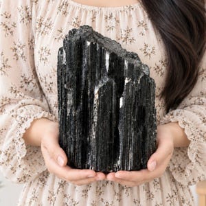 Puede incluir: Un gran espécimen de cristal de turmalina negra en bruto que presenta una estructura columnar vertical profundamente estriada con una superficie brillante y reflectante y bordes dentados e irregulares en la parte superior, de aspecto oscuro y opaco.
