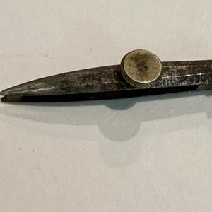 May include: Close-up of a vintage metal compass, showing the pointed tips and adjustment wheel. The compass has a worn, aged appearance with signs of rust, suggesting it's an antique drafting tool. The background is a plain, off-white surface.