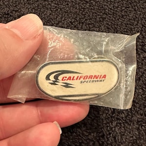 May include: A white oval-shaped pin with a black and white checkered flag design and the text "CALIFORNIA SPEEDWAY" in red letters.