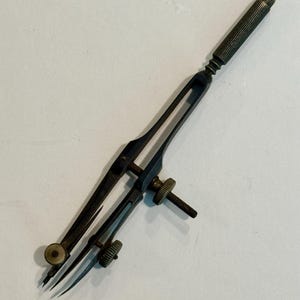 May include: Antique brass-toned drafting compass with a ribbed handle and adjustable screw mechanism. The compass features two pointed legs and a small wheel for fine adjustments. A vintage tool for technical drawing.