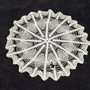 May include: A cream-colored, crocheted doily with a scalloped edge. The circular design features a detailed, open-weave pattern radiating from the center. The doily is approximately 12 inches in diameter, perfect for adding a vintage touch to any space.