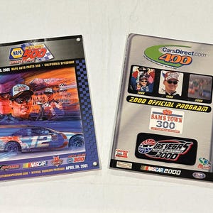 May include: Two NASCAR race program souvenir pamphlets. The left pamphlet is blue and white with a picture of a race car and drivers. The right pamphlet is silver and black with a picture of a race car and the text "CarsDirect.com 400".