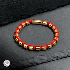 Pode incluir: Uma pulseira com um cordão vermelho vibrante entrelaçado com contas cilíndricas douradas polidas. O design inclui um fecho de metal dourado liso e alongado que prende a banda circular, criando um padrão consistente de detalhes.