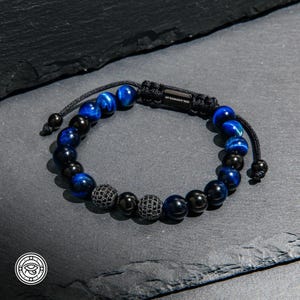 Op de afbeelding: Een kralenarmband met afwisselend blauwe en zwarte gepolijste kralen. Twee zwarte kralen zijn ingelegd met kleine zwarte steentjes. De armband heeft een zwarte koordsluiting met een rechthoekig metalen plaatje waarop My Harmony Tree is gegraveerd.