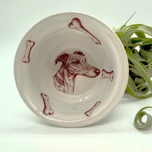 May include: A cream-colored ceramic dog bowl featuring a detailed illustration of a dog's head in reddish-brown. The bowl is decorated with bone shapes in the same color. A stylish and functional pet accessory.