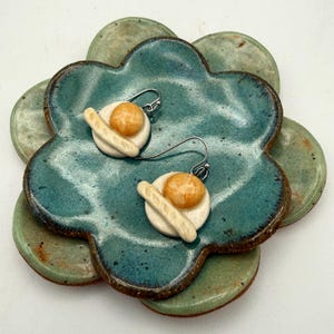 May include: A pair of handmade ceramic earrings shaped like fried eggs with a baguette. The earrings have a white base, a yellow yolk, and a beige baguette. They are displayed on a teal and green ceramic flower-shaped dish.