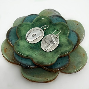May include: A pair of silver earrings with unique abstract designs. The earrings are displayed on a green and blue ceramic dish.