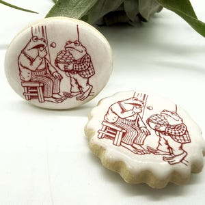 May include: Two ceramic cabinet knobs and a cookie with a whimsical frog design. The frogs are depicted in a vintage style, one seated and the other holding a basket. The knobs are round, while the cookie has a scalloped edge.