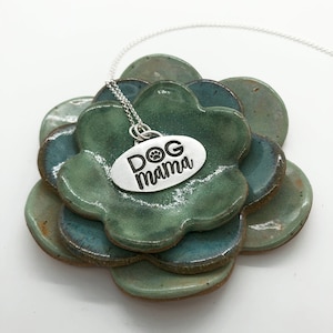 May include: Silver necklace with an oval charm that says "DOG mama" with a paw print symbol. The necklace is on a stack of green ceramic dishes.