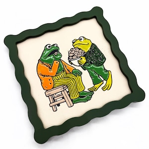 May include: A square wall art piece with a dark green wavy frame featuring a colorful illustration of two anthropomorphic frogs. One frog sits on a wooden stool wearing an orange jacket and yellow striped pants, while the other stands holding a bowl.