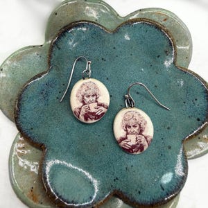 May include: A pair of oval-shaped dangle earrings featuring a sepia-toned illustration of a woman with short curly hair holding a mug. The earrings have silver-toned metal hooks and are crafted from a smooth, off-white material with a matte finish.