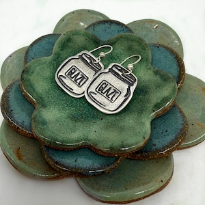 May include: A pair of silver earrings shaped like mason jars with the word "GLAZE" written on them. The earrings are displayed on a green and blue ceramic plate.