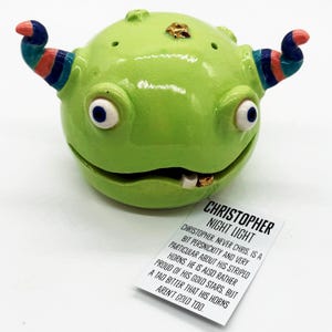 May include: A whimsical, green ceramic night light shaped like a creature with blue-eyed eyeballs, striped horns, and a tooth. The night light has the text "CHRISTOPHER NIGHT LIGHT" on a tag.