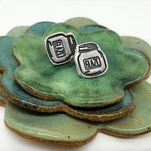 May include: A pair of silver earrings shaped like mason jars with the word "GIMT" engraved on them. The earrings are displayed on a green ceramic dish.