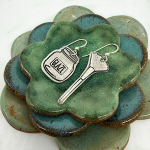 May include: A pair of silver earrings with a glaze jar and a makeup brush design. The earrings are on a green and blue ceramic plate.