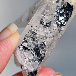 Può includere: Un cristallo di quarzo trasparente con inclusioni nere. Il cristallo è tenuto in mano da una persona. Il quarzo ha un aspetto lucido e trasparente, con depositi minerali scuri e riflettenti. È una gemma naturale e grezza.