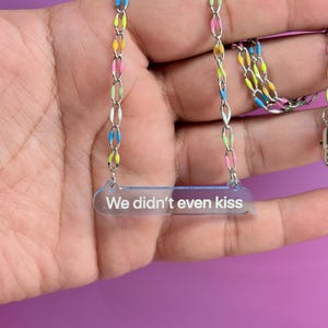 May include: A silver chain necklace with colorful enamel links in yellow, pink, blue, and white. The necklace features a clear acrylic pendant with the text "We didn't even kiss".
